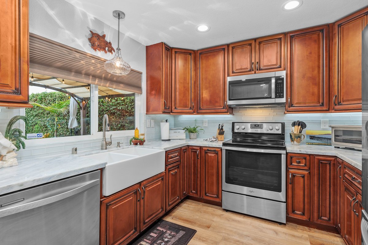 Kitchen with wooden cabinets, stainless steel stove and microwave, white farmhouse sink, dishwasher, and decorative accents, viewed through a window showing a backyard with greenery.