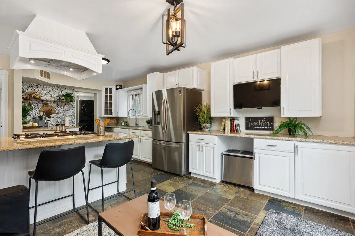 Modern kitchen with white cabinets, stainless steel refrigerator, black TV on wall, island with black chairs, slate tile flooring, decorative plants, and a wall-mounted light fixture.