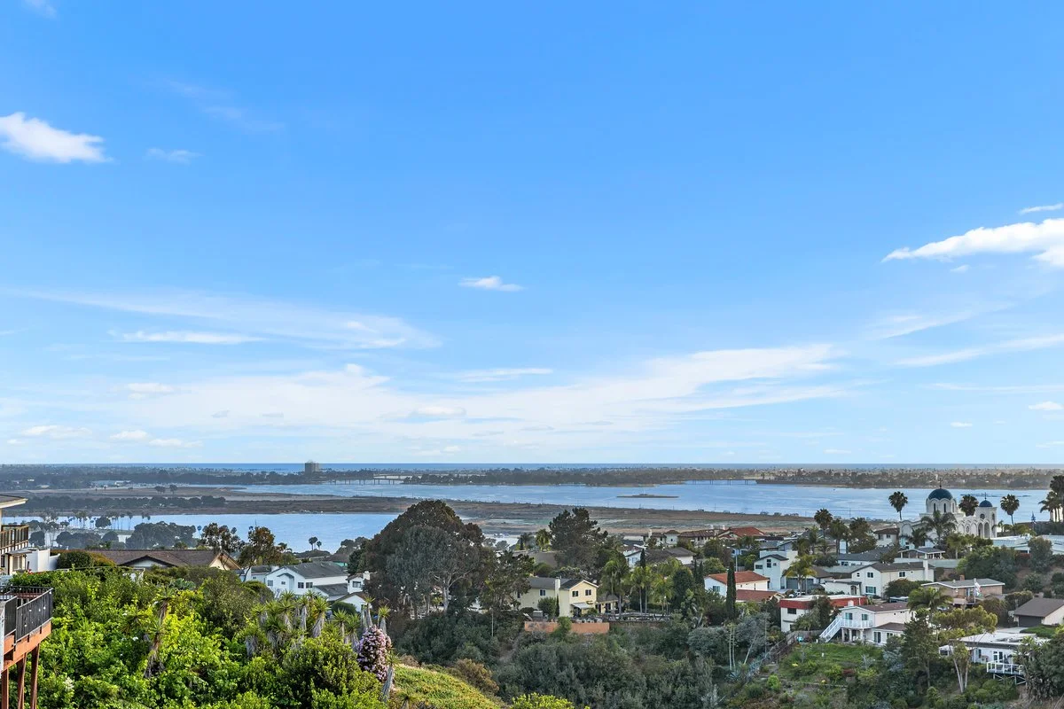 A scenic view of a coastal cityscape with houses, trees, and water under a bright blue sky with scattered clouds.