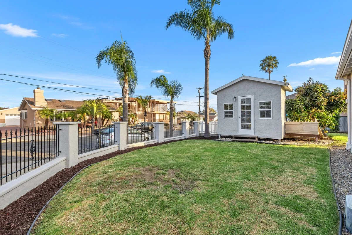 A backyard with a small white building, palm trees, a grassy lawn, a black metal fence, and neighboring houses under a blue sky.
