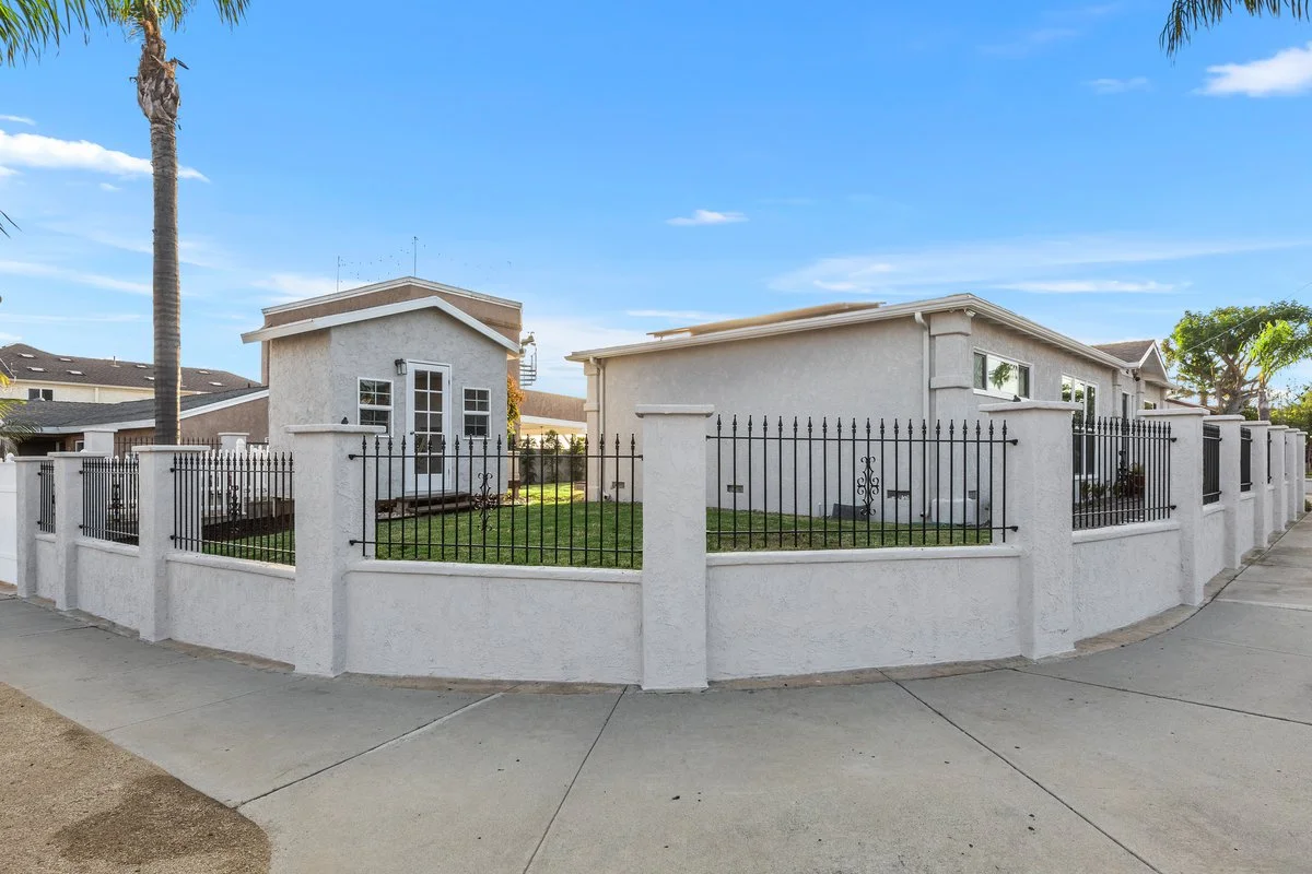 A white stucco house with a black iron fence and a small grass yard, with a palm tree on the sidewalk and a clear blue sky.
