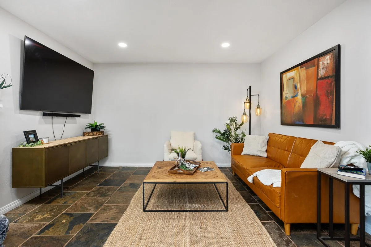 Living room with a mounted flat-screen TV, a wooden sideboard, a white armchair, a tan leather sofa, wall art, potted plants, a coffee table, and decorative lighting.