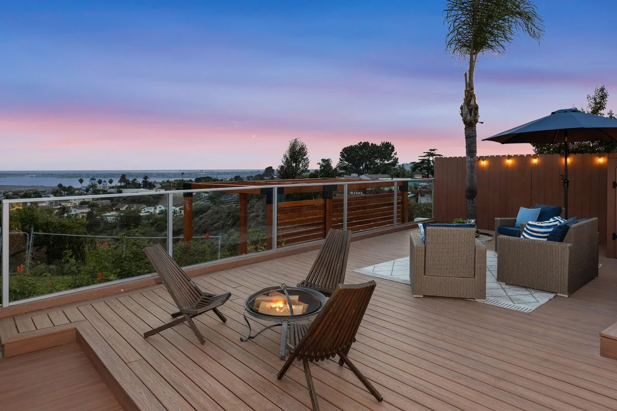 Outdoor deck with seating area, including wicker chairs, a fire pit table, an umbrella, and a view of the ocean at sunset, with trees and houses in the distance.