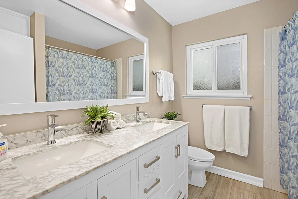 Bathroom with a double vanity with a marble countertop, a large mirror, a plant, and toiletries. There are two small frosted windows, white towels on a rack, and a shower with a patterned blue curtain.