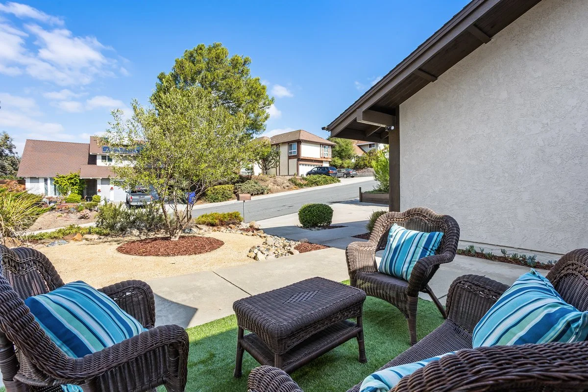 Outdoor patio area with wicker chairs and striped cushions, adjacent to a house with a beige wall, overlooking a landscaped yard with trees and bushes, under a blue sky.
