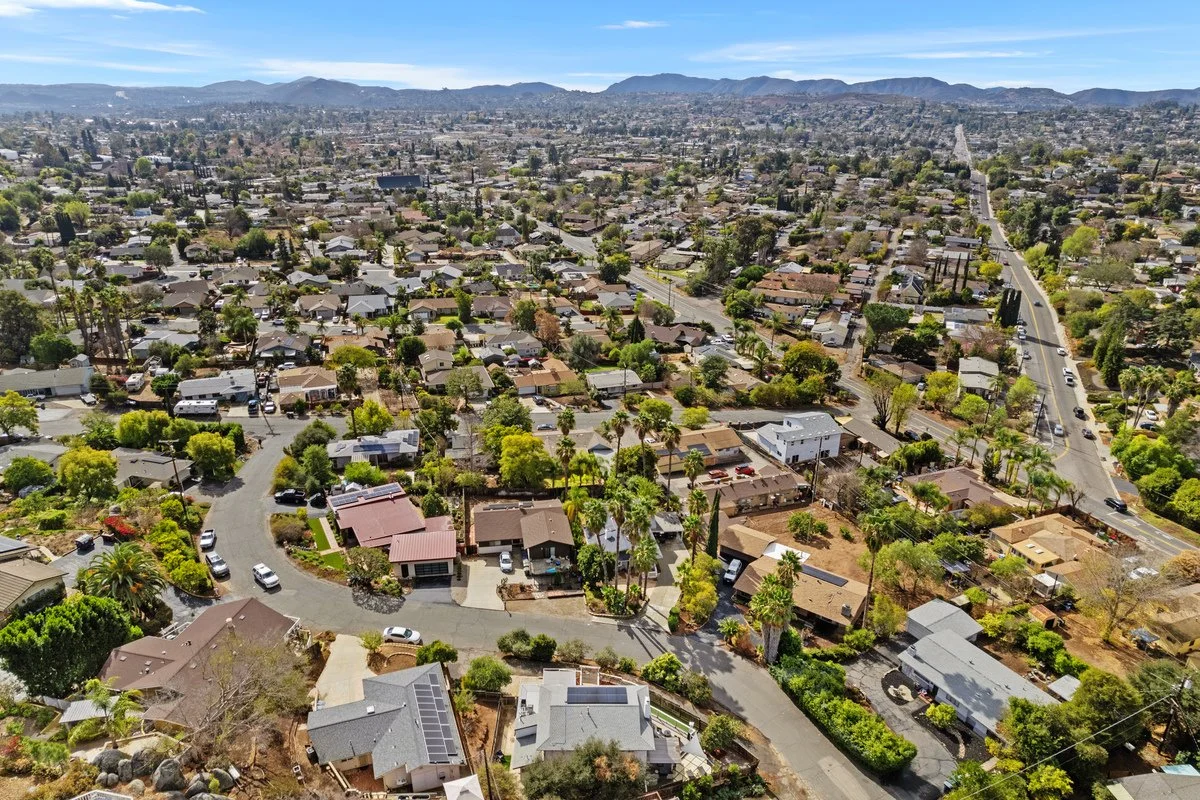 Aerial view of a suburban neighborhood with single-family homes, tree-lined streets, and a mountain range in the distance under a clear blue sky.