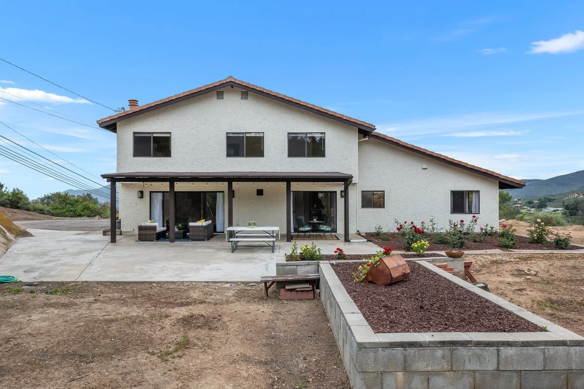 A two-story house with a stucco exterior and a tiled roof, featuring an outdoor patio with seating and a garden with flowers, set in a rural area with mountains in the background and a partly cloudy sky overhead.