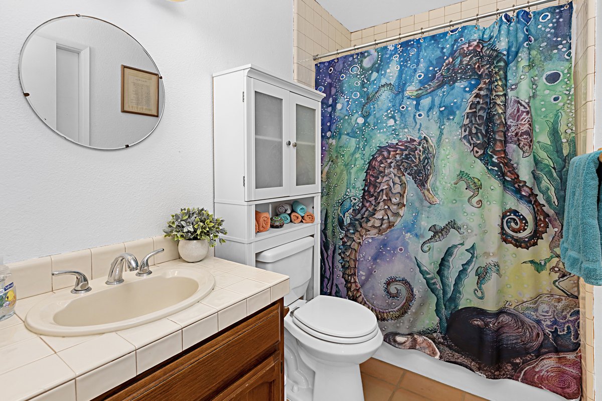 Bathroom with a seahorse-themed shower curtain, white wall, round mirror, sink with beige tiles, small white cabinet, and a blue towel.
