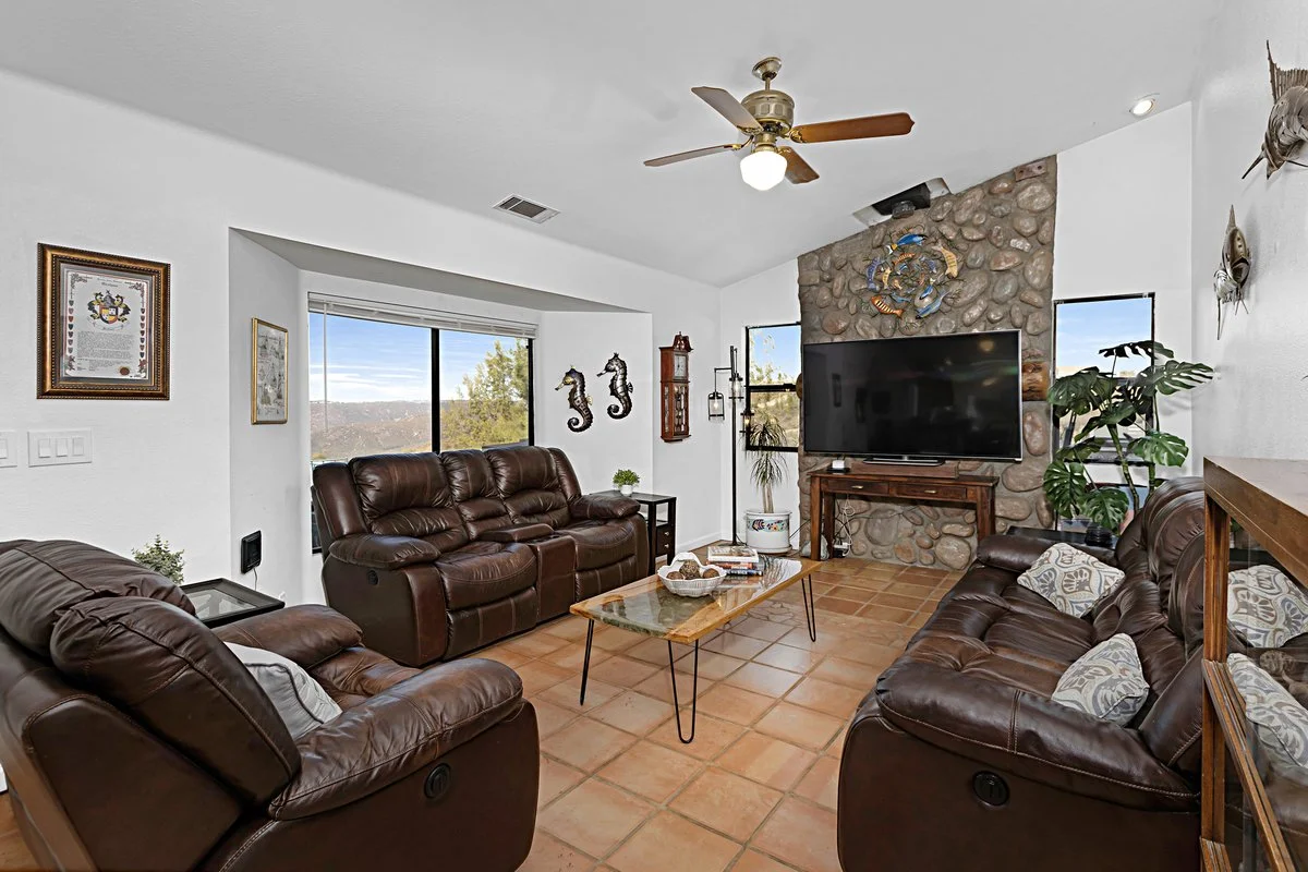 Living room with three brown leather sofas, a stone fireplace with a flat-screen TV, a ceiling fan, two windows showing a scenic outdoor view, and various decorative elements.