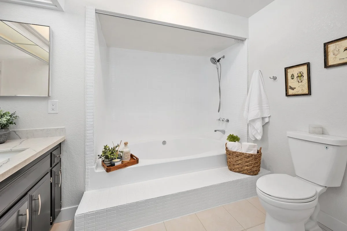 Bathroom with shower and bathtub, white walls, brown woven basket with rolled towels, white towel hanging, decorative framed pictures, gray vanity with marble countertop, and small plant on the counter.