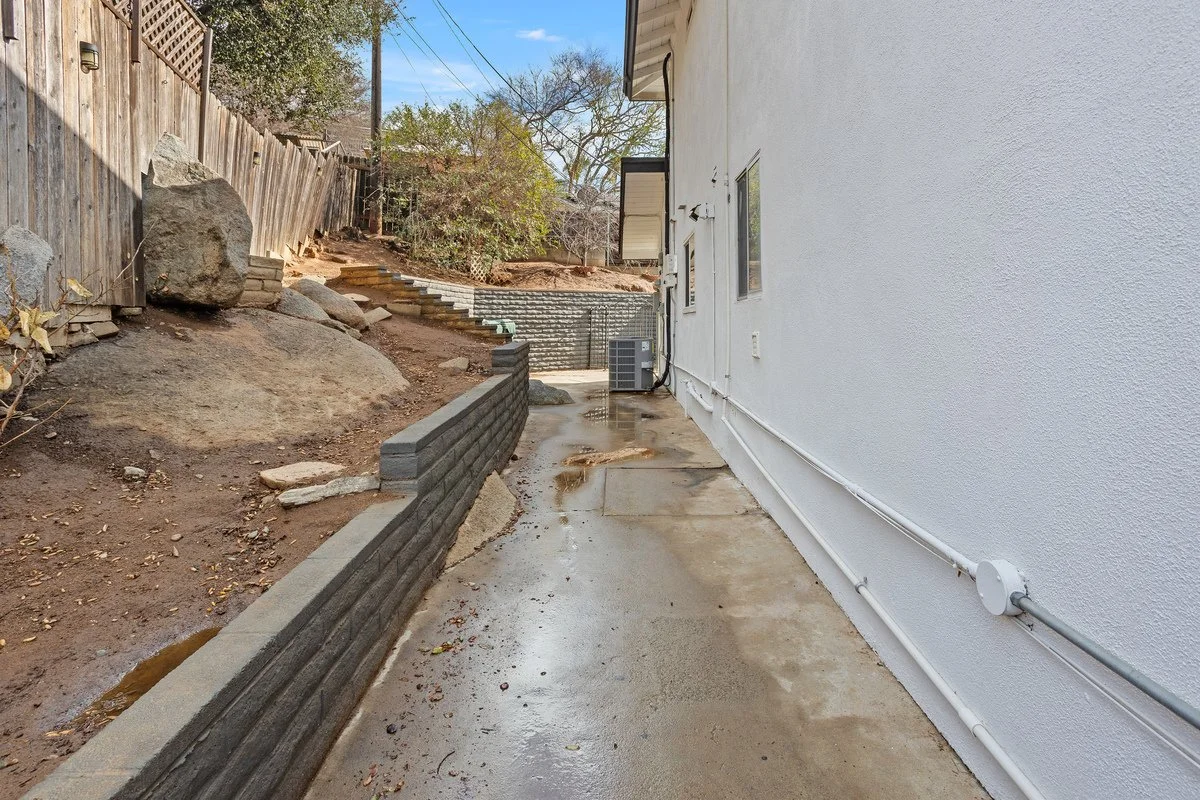Side of a house with a white exterior wall, electrical wiring and an outdoor air conditioning unit, next to a dirt hillside with rocks and trees, and a concrete walkway with water puddles.