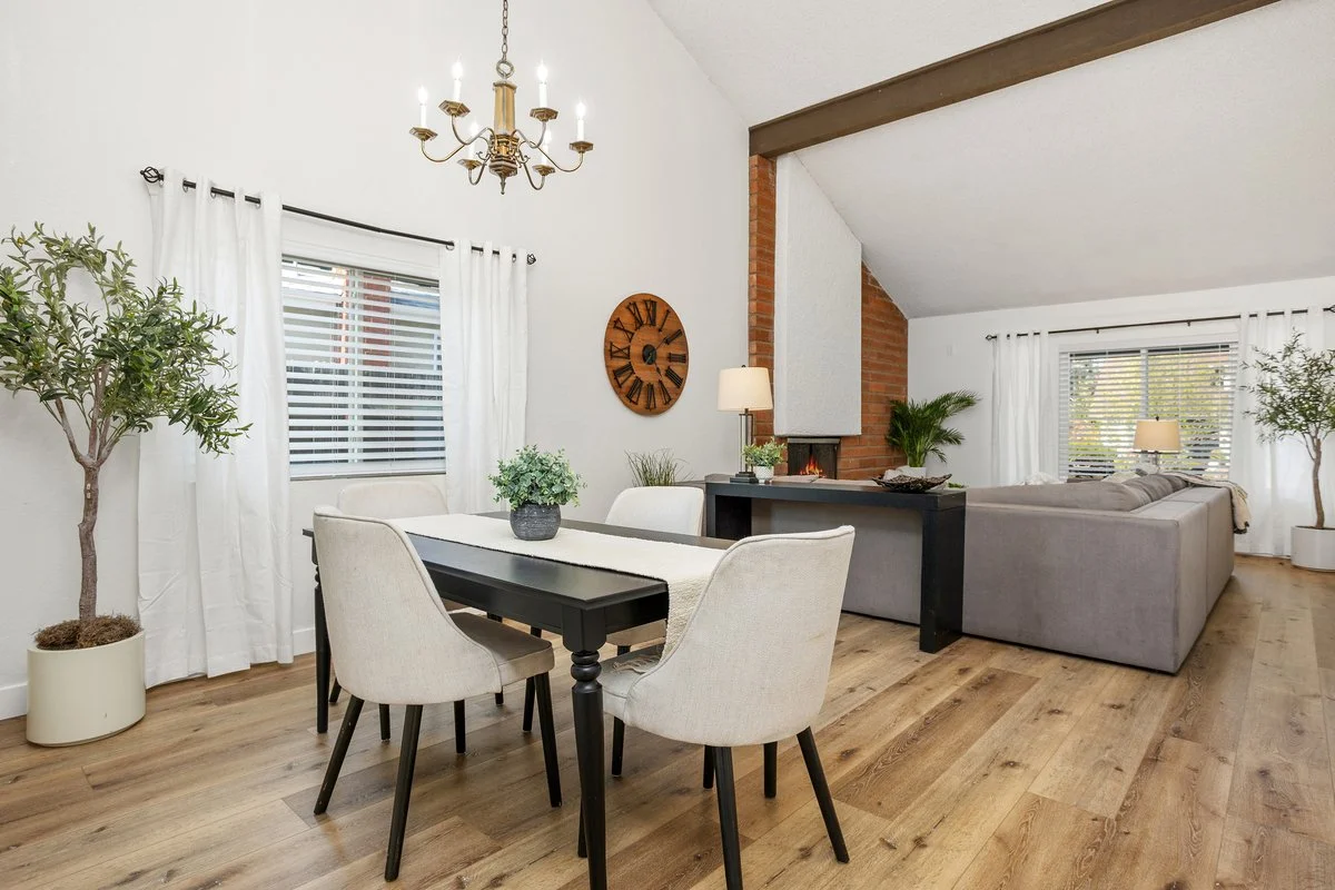 Open-concept living and dining room with a wooden dining table, beige chairs, a gray sectional sofa, a brick fireplace, and large windows with white curtains