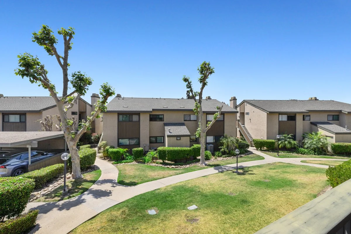 A residential apartment complex with multiple two-story buildings, surrounding landscaped lawns, trimmed bushes, trees, walkways, and parking spaces, under a clear blue sky.