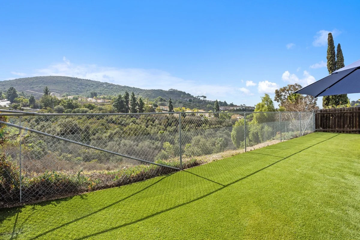 Backyard with green lawn, chain-link fence, and scenic hilly landscape in the background under a partly cloudy blue sky.