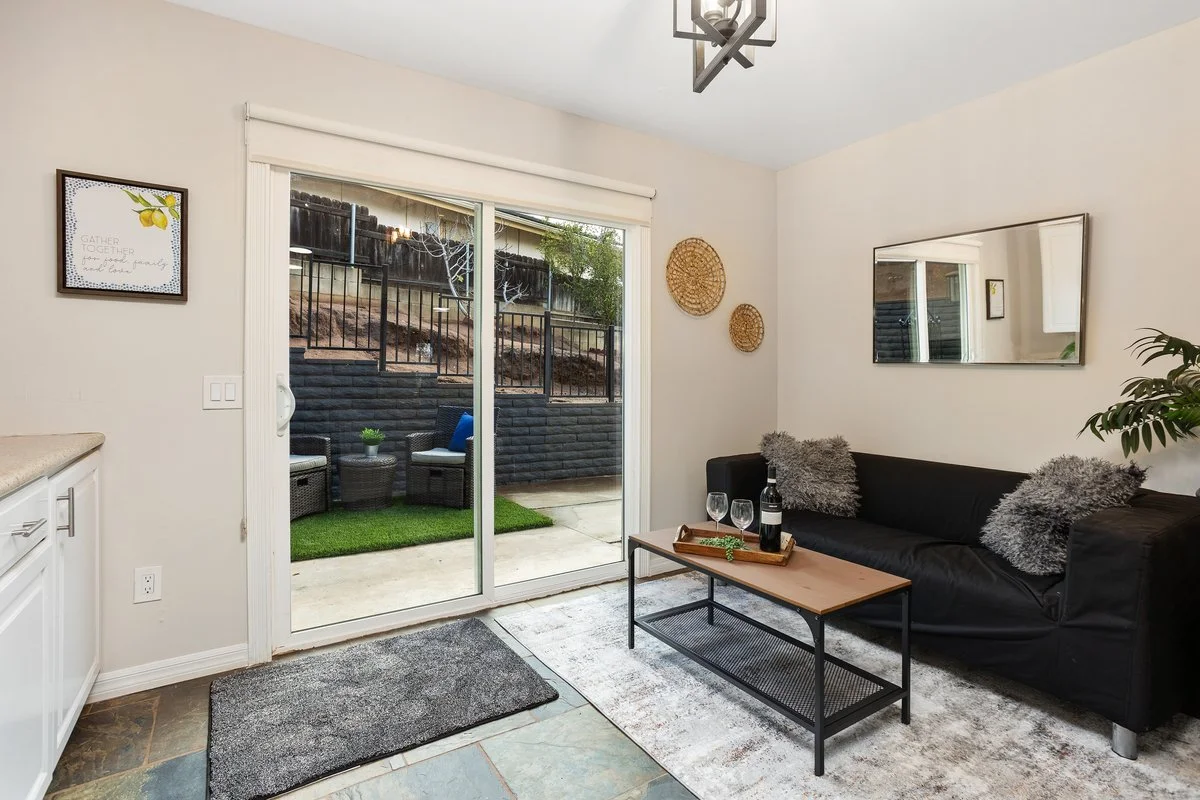 Living room with sliding glass door leading outside to a patio with patio furniture, a black sofa with gray pillows, a wall mirror, two woven wall hangings, a picture frame, a small coffee table with wine glasses and a bottle, and a sliding door with