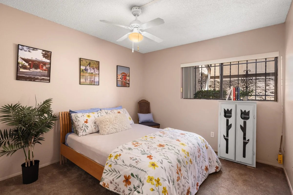 A bedroom with a bed covered in a floral duvet, a tall green plant in a black pot, framed pictures on the wall, a window with security bars, and a white cabinet with decorative cutouts on top.