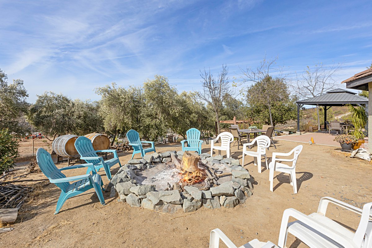 Outdoor patio area with a fire pit surrounded by colorful plastic chairs, some tables, trees, and a clear blue sky
