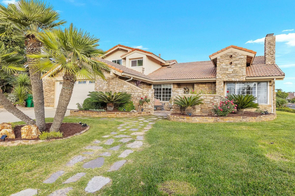 Front view of a house with a stone exterior, a tiled roof, and a well-maintained lawn with pathway and tropical plants.