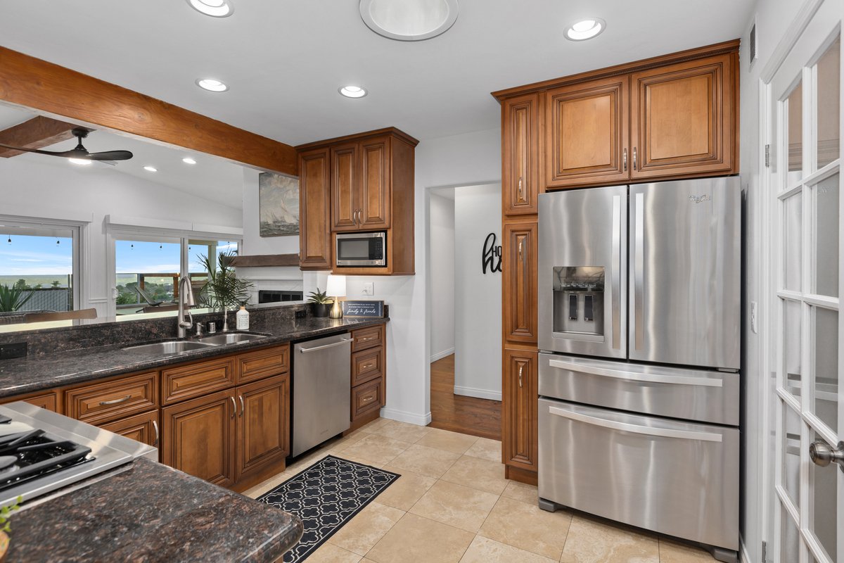 Kitchen with wooden cabinets, stainless steel refrigerator, black granite countertops, tile floor, and view of living room through opening.