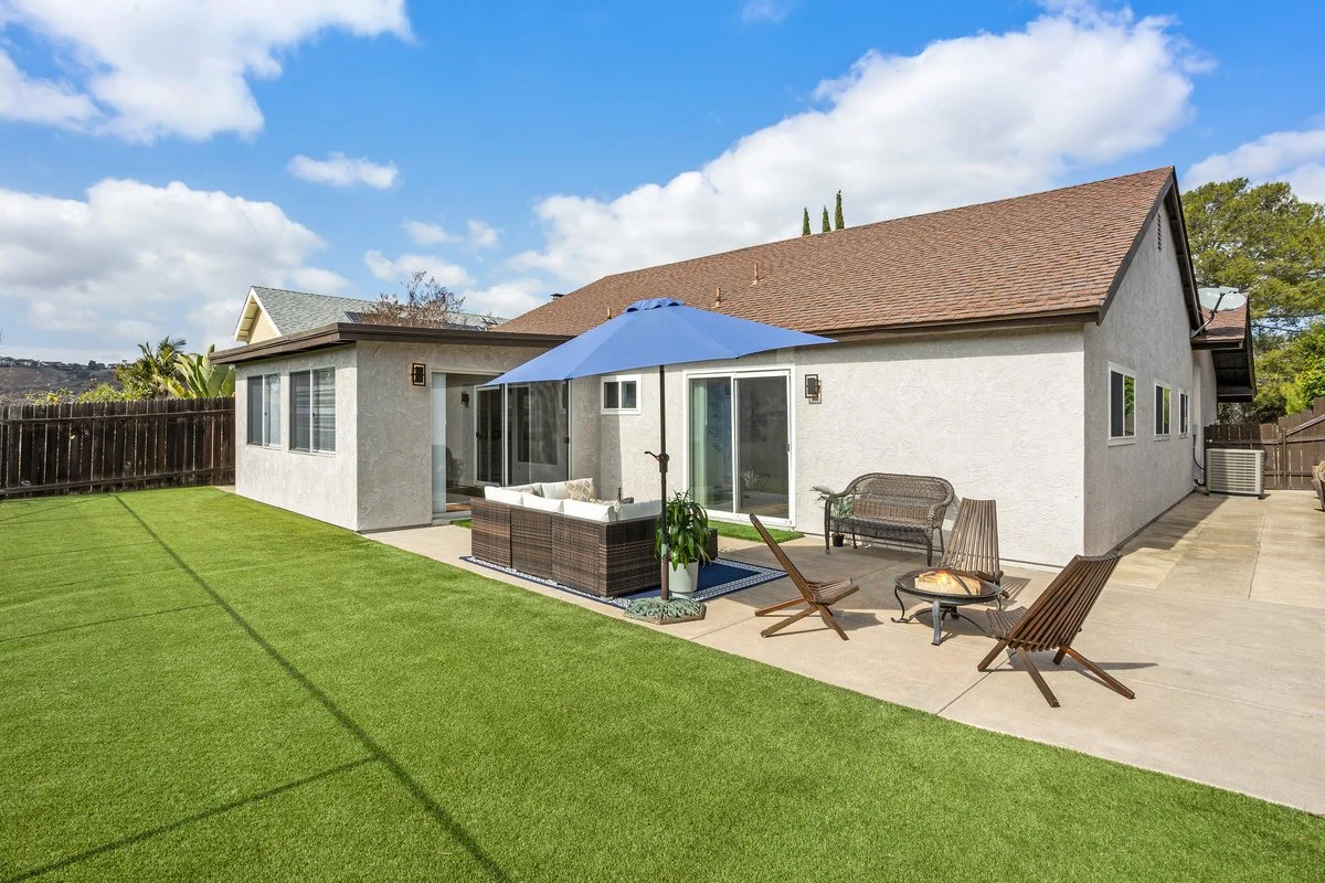 Backyard with patio furniture, a blue umbrella, and a lawn, attached to a house with sliding glass doors and multiple windows under a partly cloudy sky.