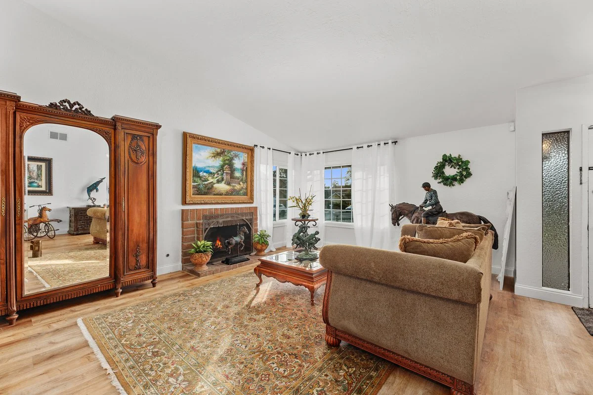 Living room with large windows, a brick fireplace, antique wooden wardrobe with mirror, Victorian-style carpet, beige sofa, and decorative figurines including a boy on a horseback and a horse sculpture.