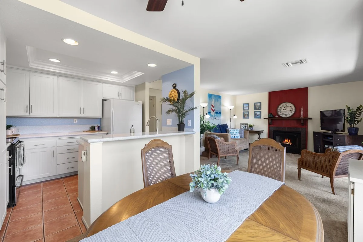 Open-concept living room and kitchen with white cabinets, a small dining table with a flower arrangement, and a living area with a fireplace and TV.