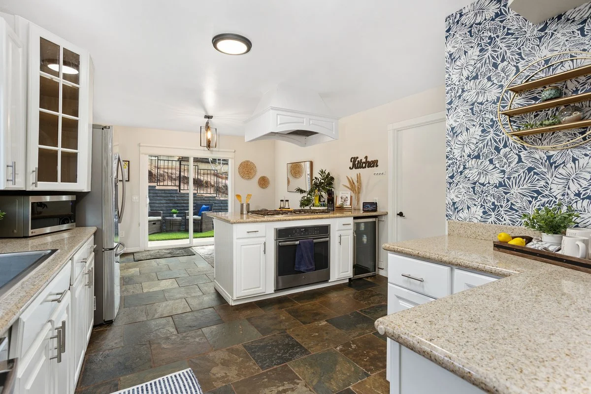 Kitchen with white cabinets, granite countertops, and a sliding glass door leading to an outdoor space with patio furniture and steps. There is a stove, microwave, mini refrigerator, and decorative items including plants and wall decor.