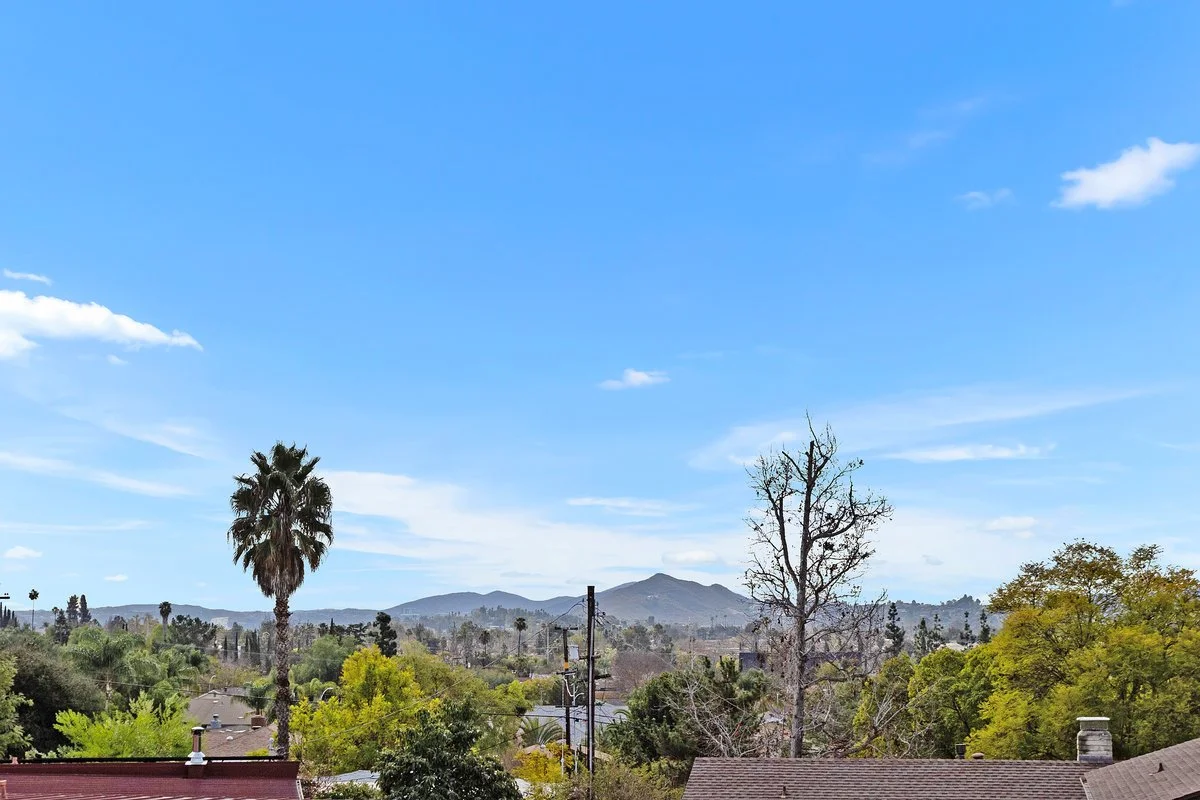 Scenic view of a suburban area with trees, hills in the background, and a clear blue sky with some clouds.
