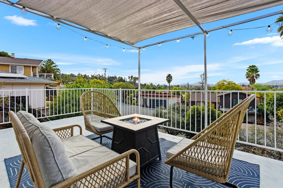 Outdoor balcony with a beige canopy, string lights, wicker chairs, a sofa, a fire pit, and a view of trees and neighborhood houses under a clear blue sky.
