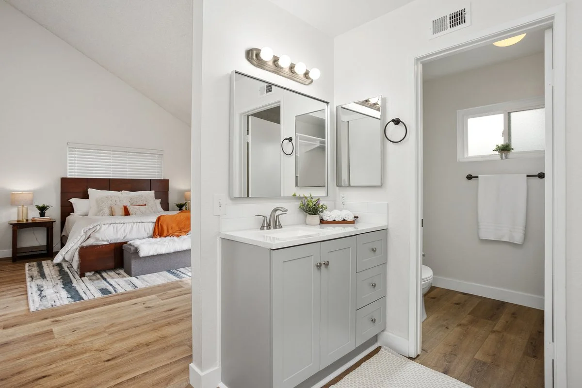 A bathroom vanity with a gray cabinet and two mirrors, adjacent to a small bathroom with a window, in a bedroom with wood flooring.
