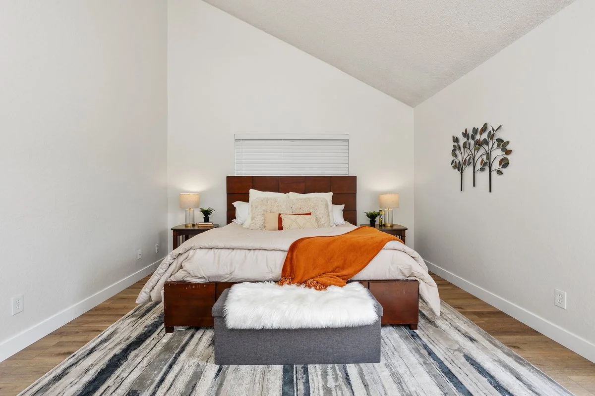 A neatly made bed with a wooden headboard, accented with white and beige pillows, and an orange blanket. There are two small bedside tables with lamps and plants. A white furry ottoman is at the foot of the bed, on a multicolored striped area rug, in