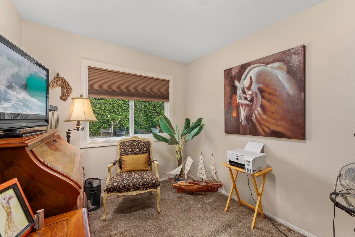 A cozy room with a window, a patterned armchair with a yellow pillow, a decorative ship model, a potted plant, a horse painting on the wall, a TV, a lamp, a printer on a wooden stand, and a fan.