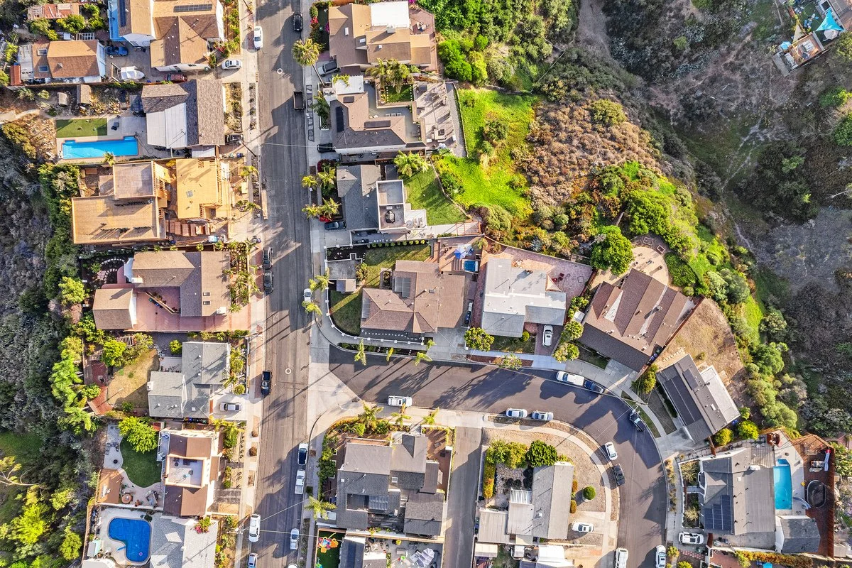 An aerial view of a residential neighborhood with houses, trees, and a winding street. Some houses have swimming pools, and there is a hillside with dense trees on one side.