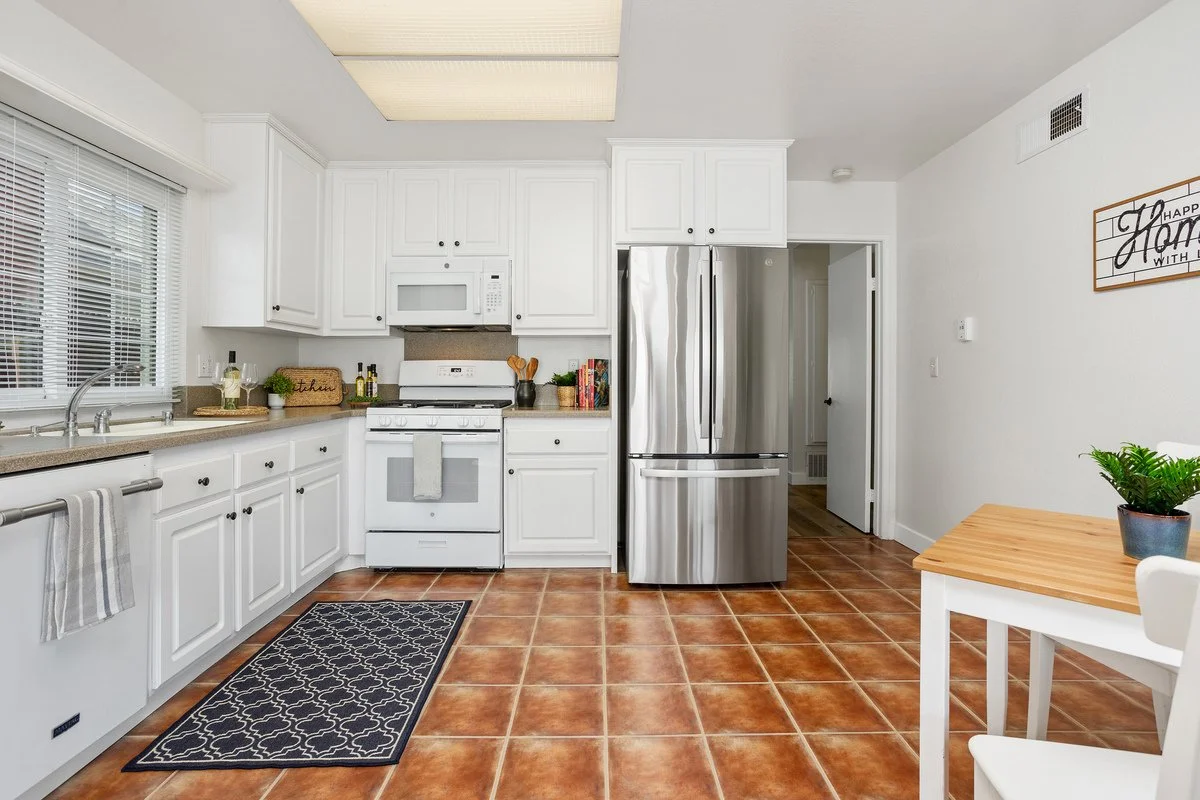 Kitchen with white cabinets, stainless steel refrigerator, white stove, microwave, tiled floor, window with blinds, and a small dining table with a plant.