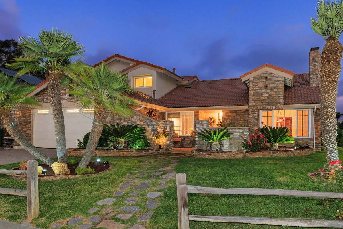 A house with a stone exterior, surrounded by palm trees, illuminated windows, a well-manicured lawn, and a stone pathway leading to the front door at dusk.