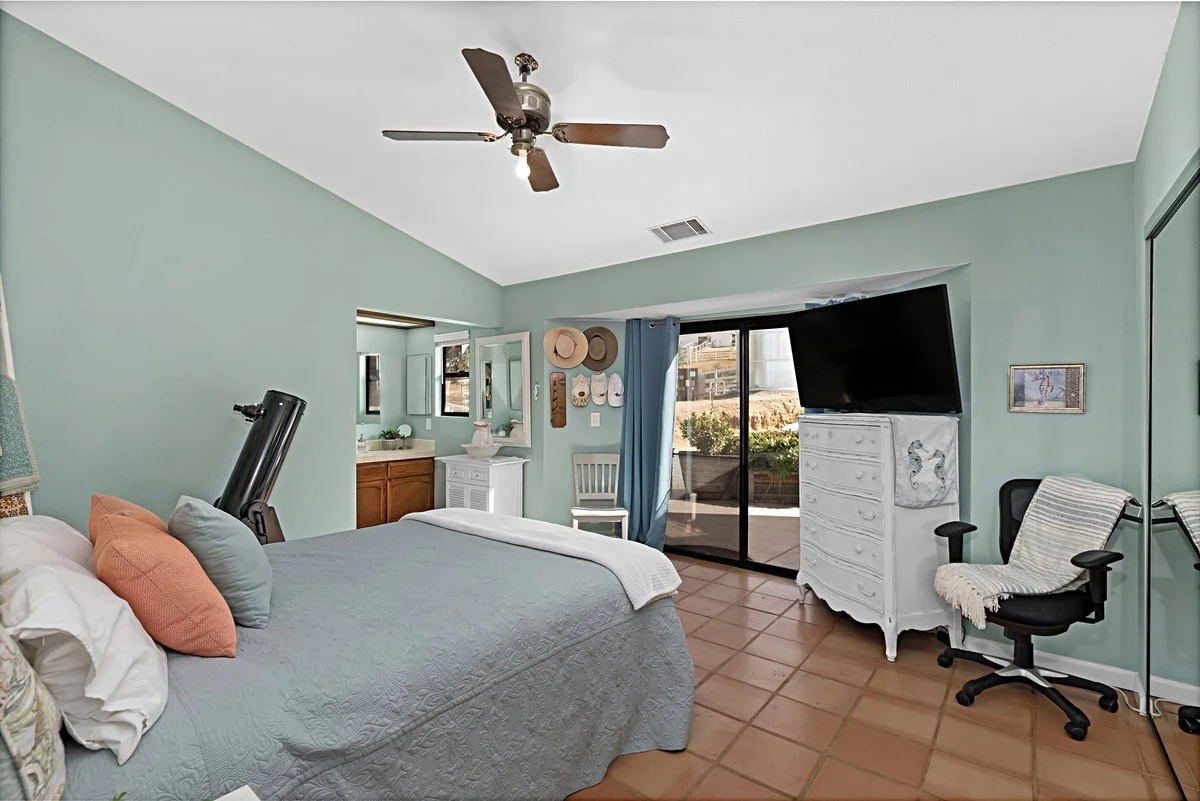 A bedroom with a bed, pillows, a ceiling fan, a dresser, a TV, a sliding glass door leading to an outdoor patio, and various decorative items.