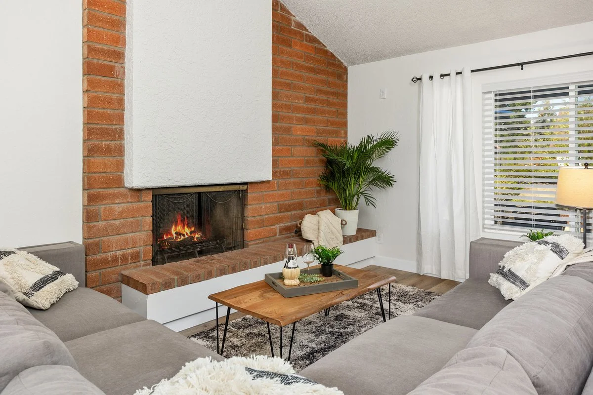 Living room with a fireplace, gray sofas, a wooden coffee table, houseplants, a window with blinds and curtains, and a rug.