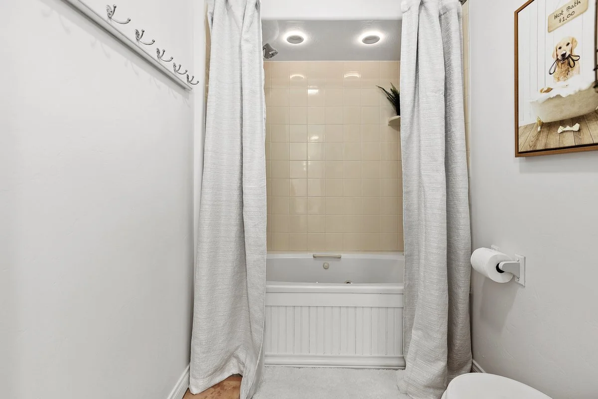 Bathroom with a shower and bathtub behind white curtains, small potted plant on the shelf, picture of a dog on the wall, and a toilet with a roll of toilet paper on the wall.