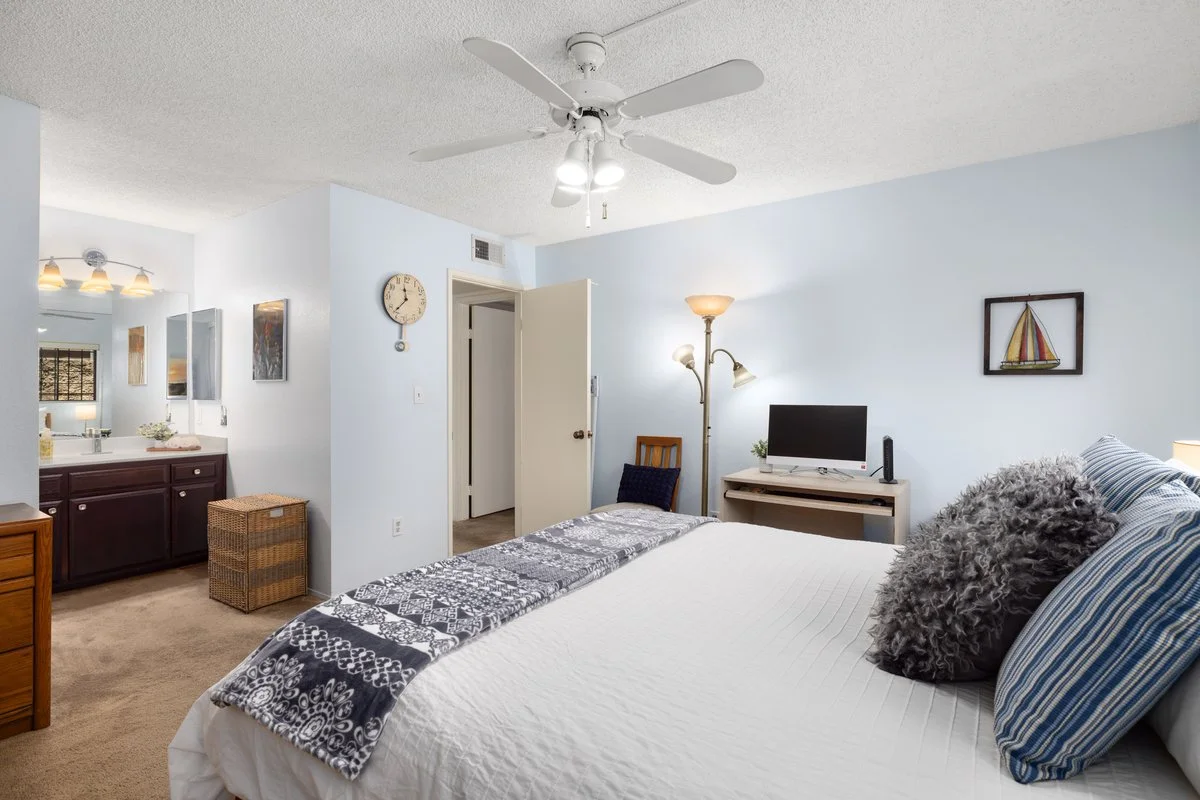 A bedroom with a white bed, fluffy pillows, a small TV, a standing floor lamp, a ceiling fan, and a view into a bathroom with a mirror and vanity.
