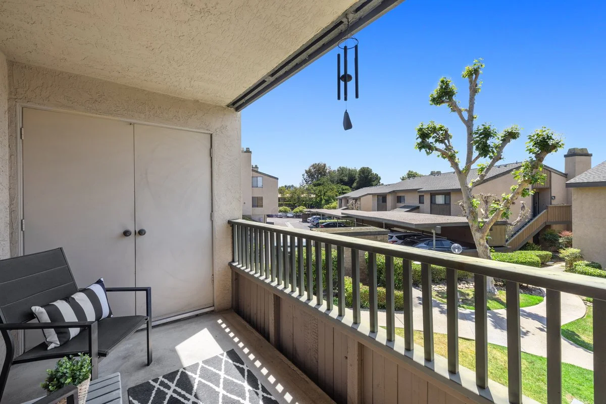 Balcony with a bench, potted plant, wind chime, and view of apartment buildings, parking lot, trees, and blue sky.