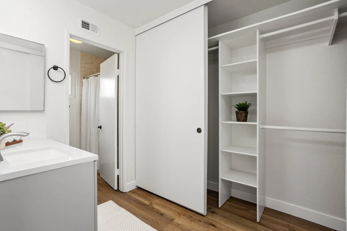 Empty white closet with sliding doors and built-in shelves in a bathroom, next to a small white sink with a plant, in a room with wood flooring.