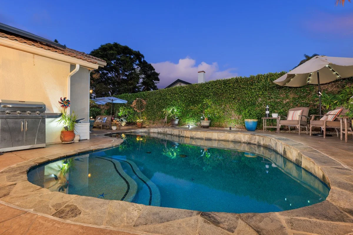 Evening view of a backyard with a swimming pool, lounge chairs, umbrellas, potted plants, and a hedge, with a house wall on the left and trees in the background.