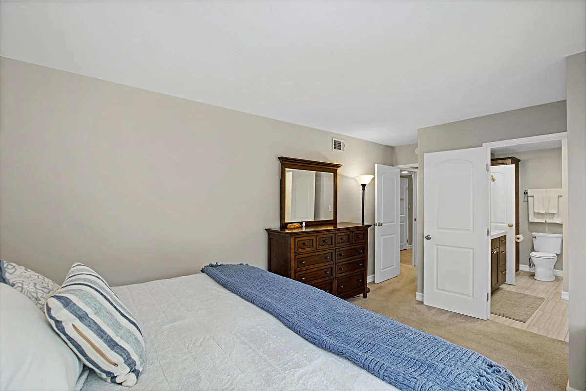 A bedroom with a bed covered in white bedding with striped pillows and a blue throw, a wooden dresser with a mirror, and an open door leading to an en-suite bathroom with a toilet and towels.