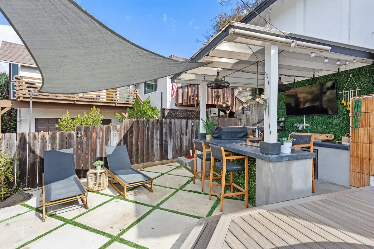 Outdoor patio area with lounge chairs, bar seating, string lights, a large flat-screen TV, and decorative greenery.