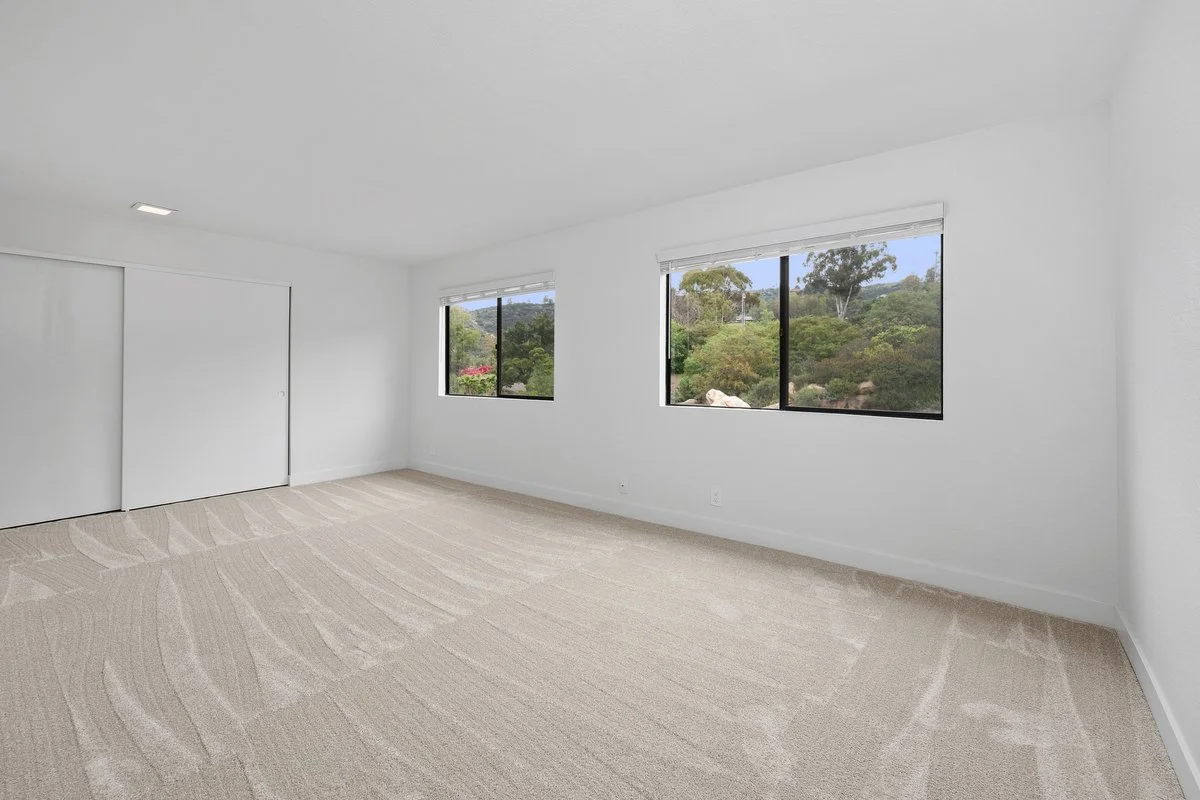 Empty room with white walls, beige carpet, and three large windows showing trees and greenery outside.