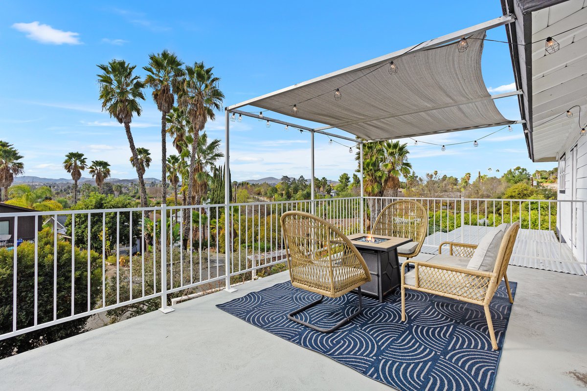 Outdoor balcony with four wicker chairs, a small table with a fire pit, and string lights, overlooking palm trees and a bright blue sky.