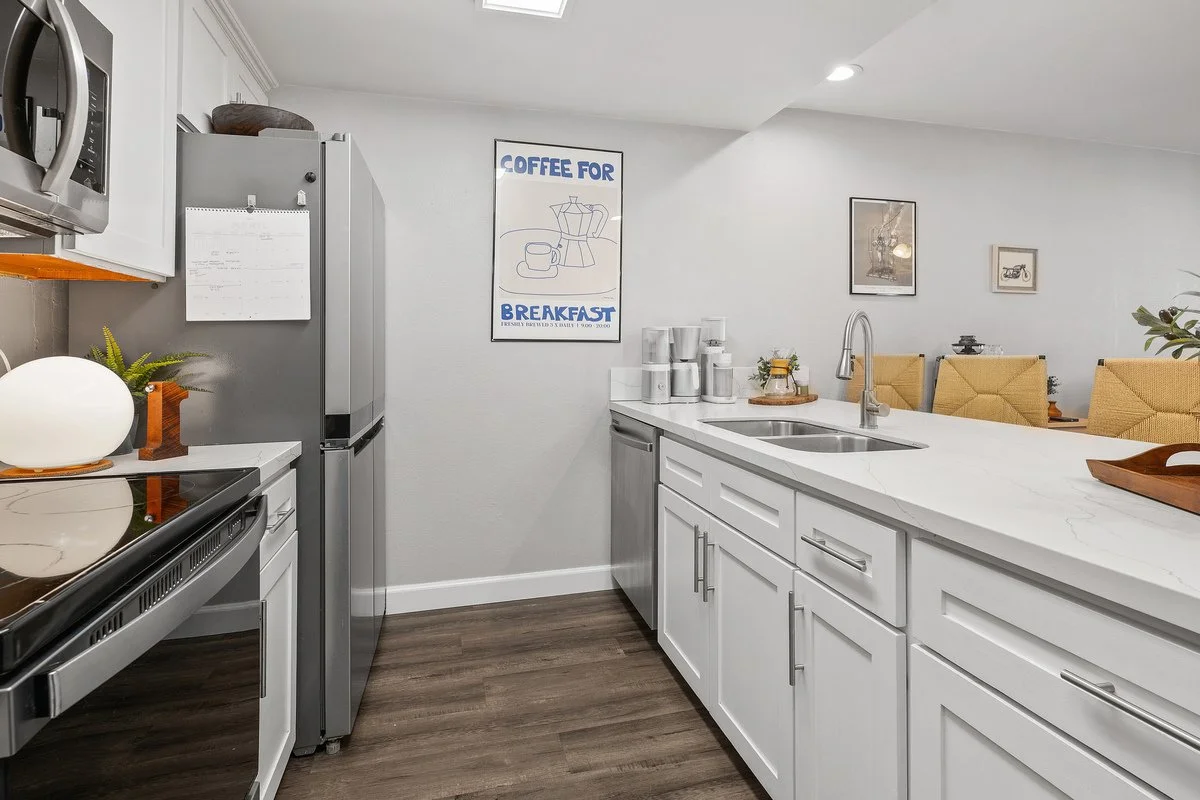 Modern kitchen with white cabinets, stainless steel appliances, and a white marble countertop. Decorative plants and framed art are on the wall, with a kitchen island and bar chairs visible.