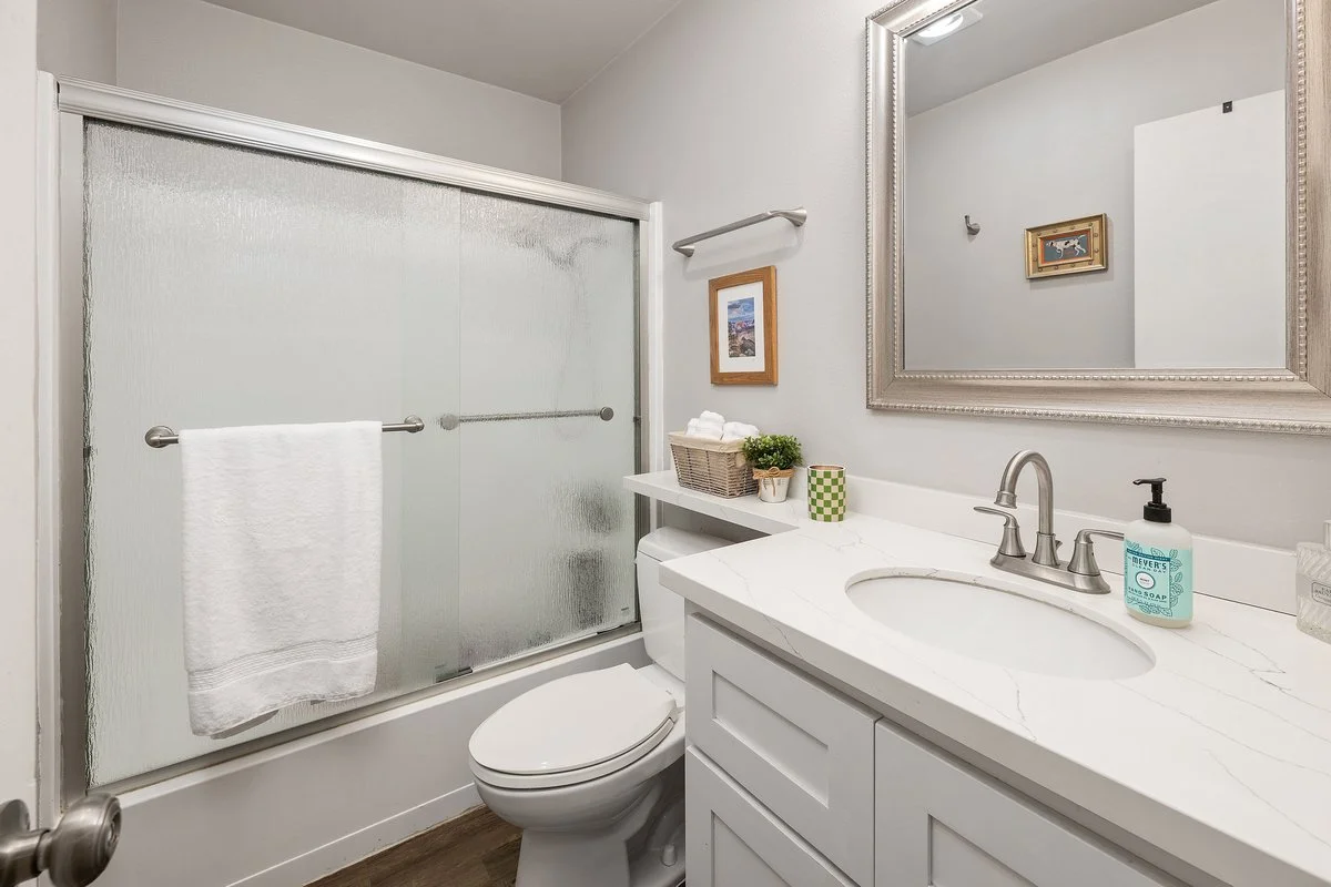 Bathroom with a glass sliding shower door, white toilet, white vanity with a marble countertop, large framed mirror, soap dispenser, small framed pictures, towel rack, and decor items.