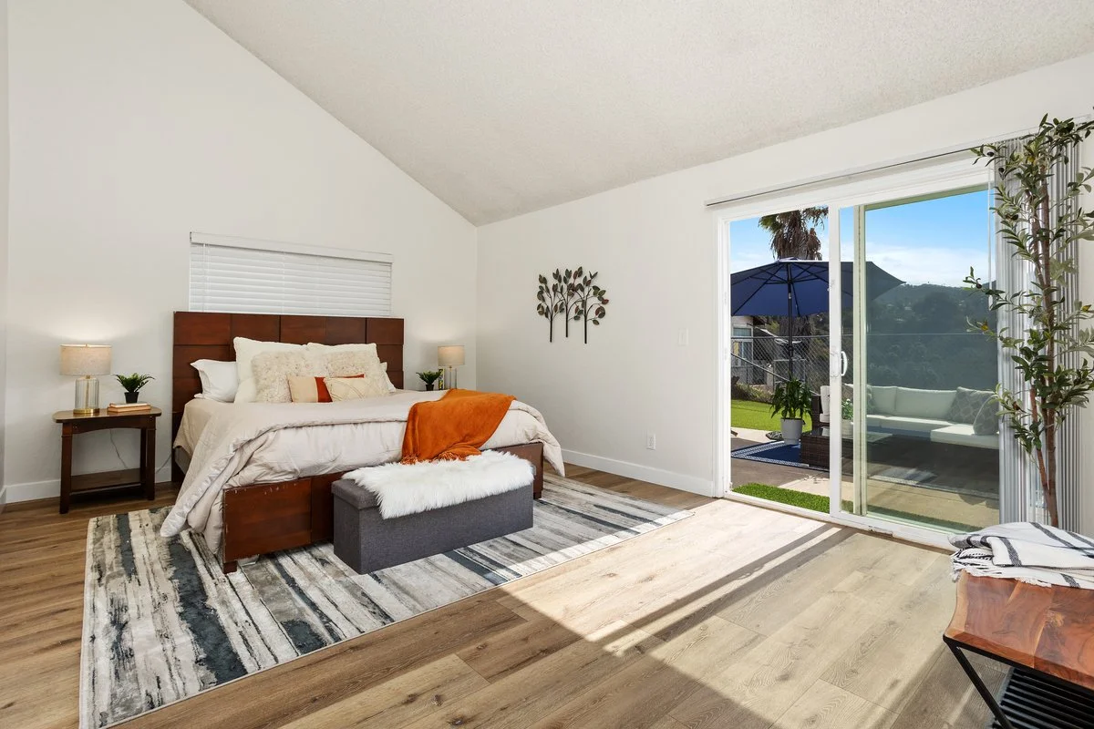 Bedroom with a wooden bed frame, white bedding, orange throw blanket, and bedside lamps, with sliding glass doors leading to an outdoor patio with a seating area and umbrella.
