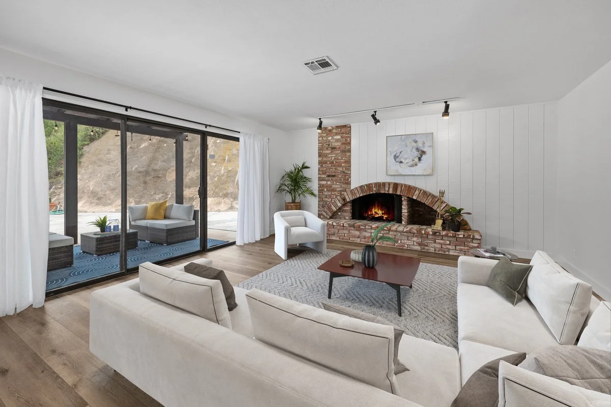 Living room with white sofas, a brick fireplace with a fire, a side chair, a coffee table with vases and decor, sliding glass doors leading to a patio with outdoor furniture, and a hillside outside.
